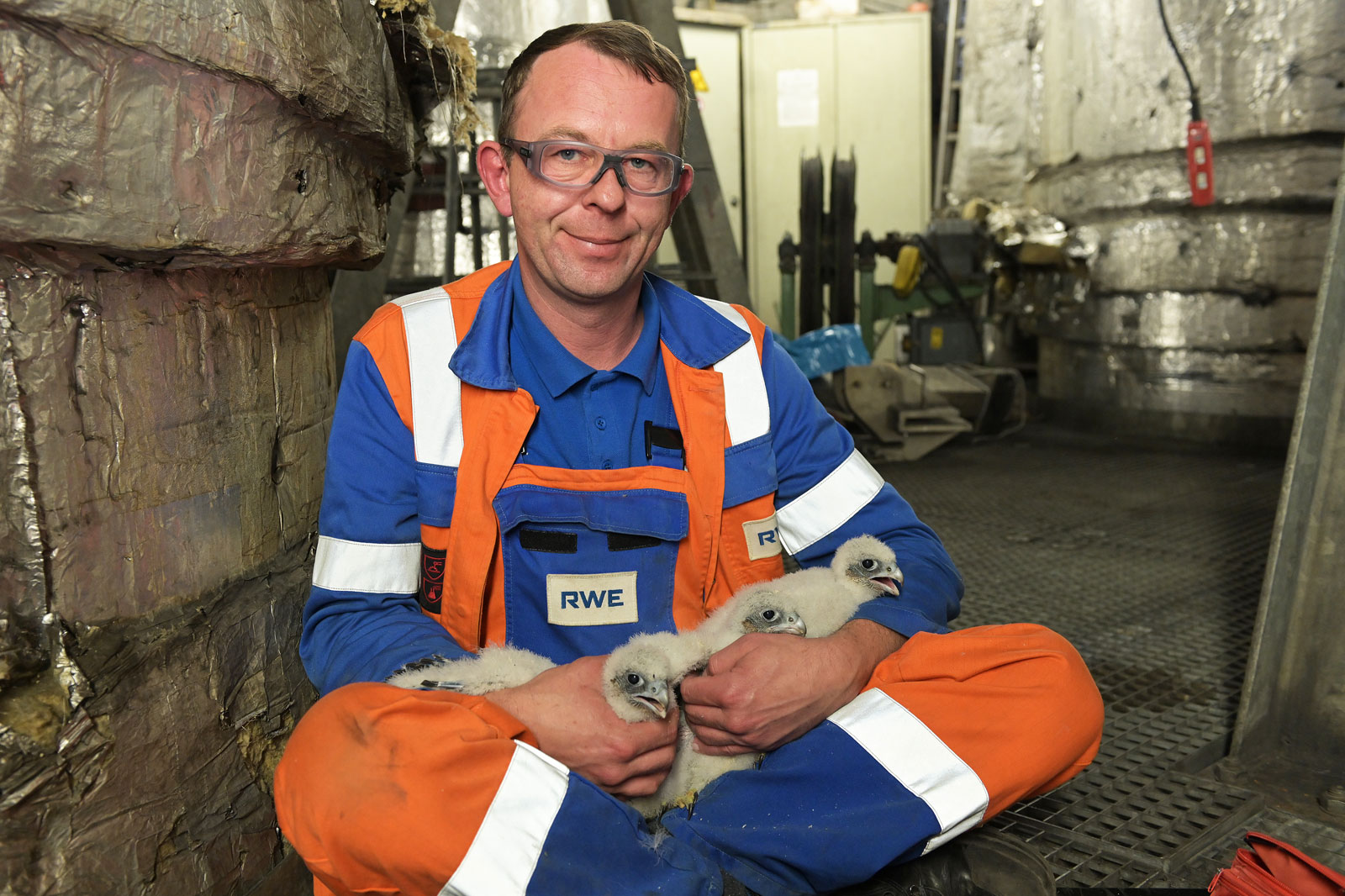 Eine Person in einer orange-blauen Uniform hält drei flauschige Küken in einer schwach beleuchteten Industrieumgebung.