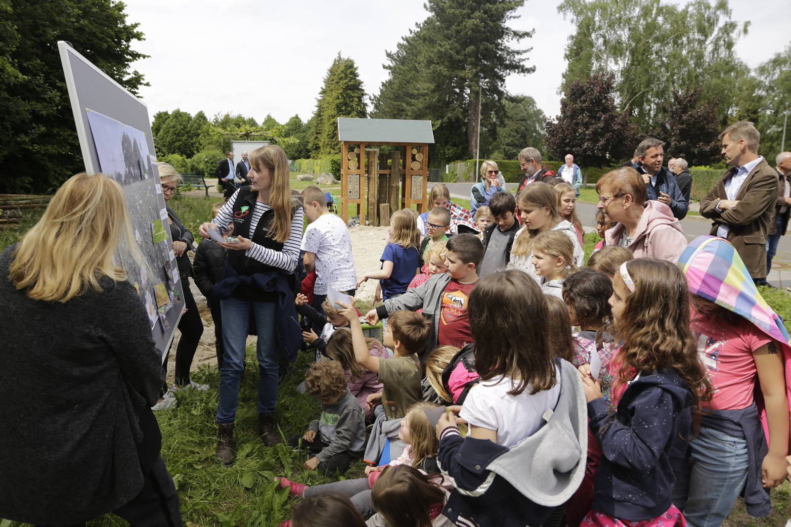 Eine Gruppe von Kindern versammelt sich im Freien um ein Informationsbrett und beteiligt sich an einer Lernaktivität mit einem Erwachsenen.