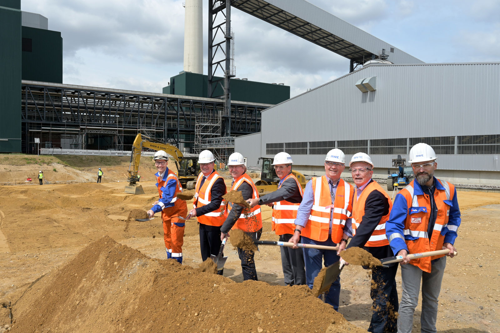 Eine Gruppe von Bauarbeitern in Sicherheitskleidung, die Erde an einer Baustelle mit Maschinen im Hintergrund schaufeln.