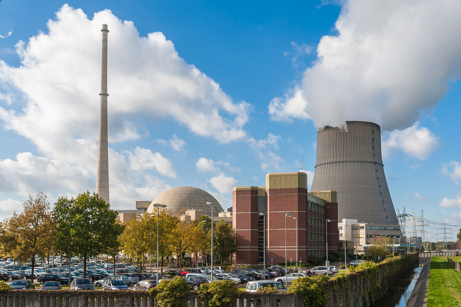 Ein Kernkraftwerk mit Kühltürmen, einem hohen Schornstein und einem Parkplatz, umgeben von Bäumen unter einem blauen Himmel.