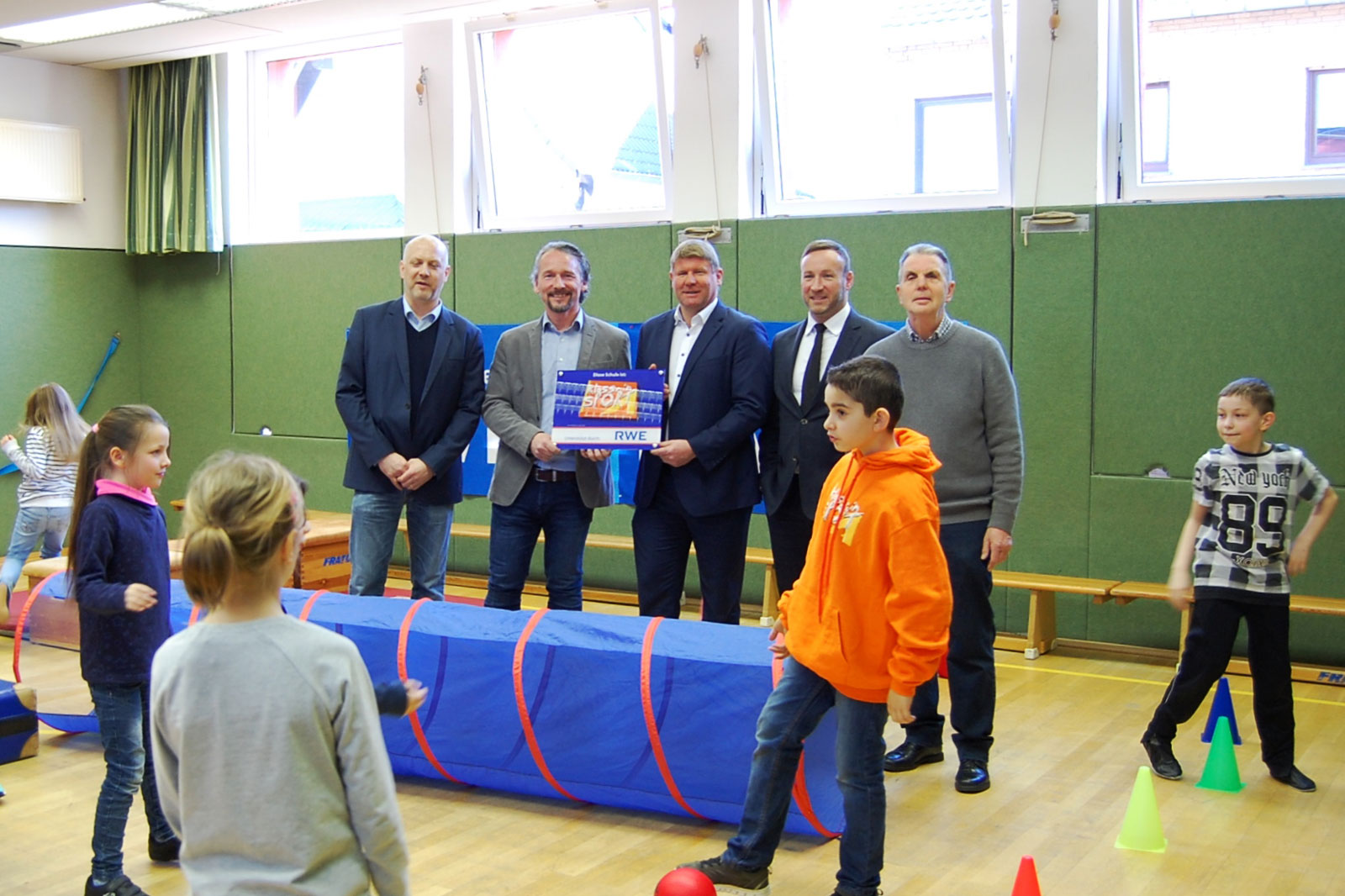 Eine Gruppe von Erwachsenen und Kindern in einer Turnhalle, die mit Sportgeräten und einem blauen Tunnel aktiv sind.