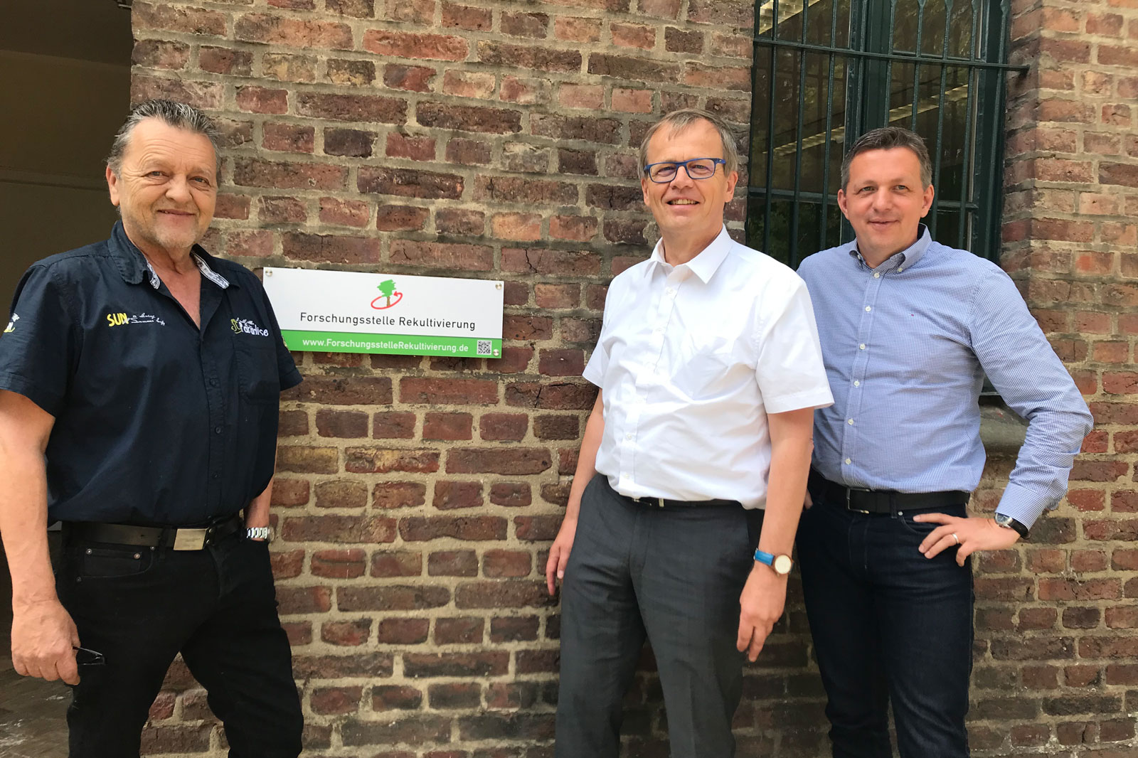 Drei Männer stehen vor einer Backsteinmauer mit einem Schild der Forschungstelle Rekultivierung.