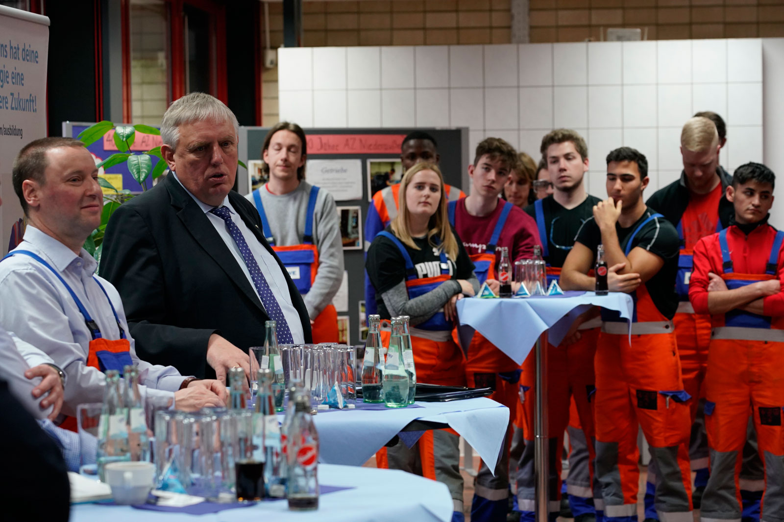 Eine Gruppe von Personen in orangefarbigen Overalls versammelt sich an einem Tisch mit Getränken und einem Präsentationshintergrund.