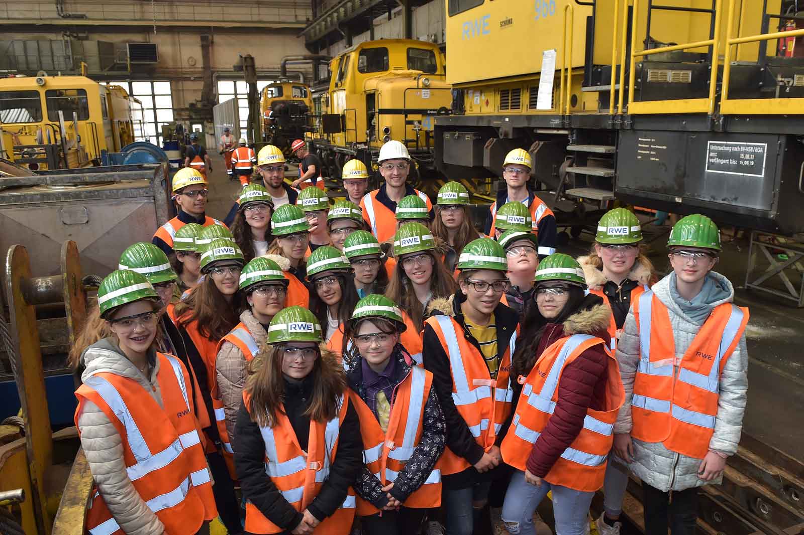 Eine Gruppe von Personen trägt Sicherheitswesten und Helme und steht in einer Werkstatt voller Maschinen.