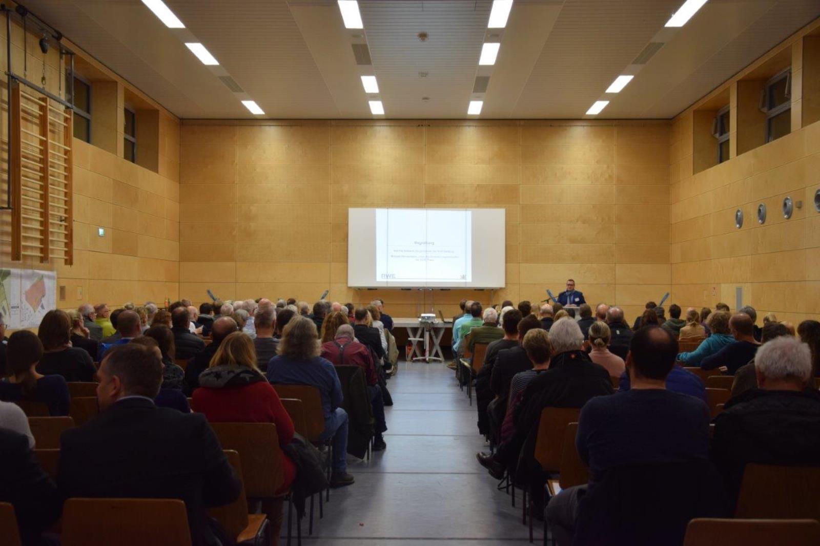 Ein großes Publikum sitzt in einem Saal und hört aufmerksam einem Referenten zu, der Informationen auf einem Bildschirm präsentiert.