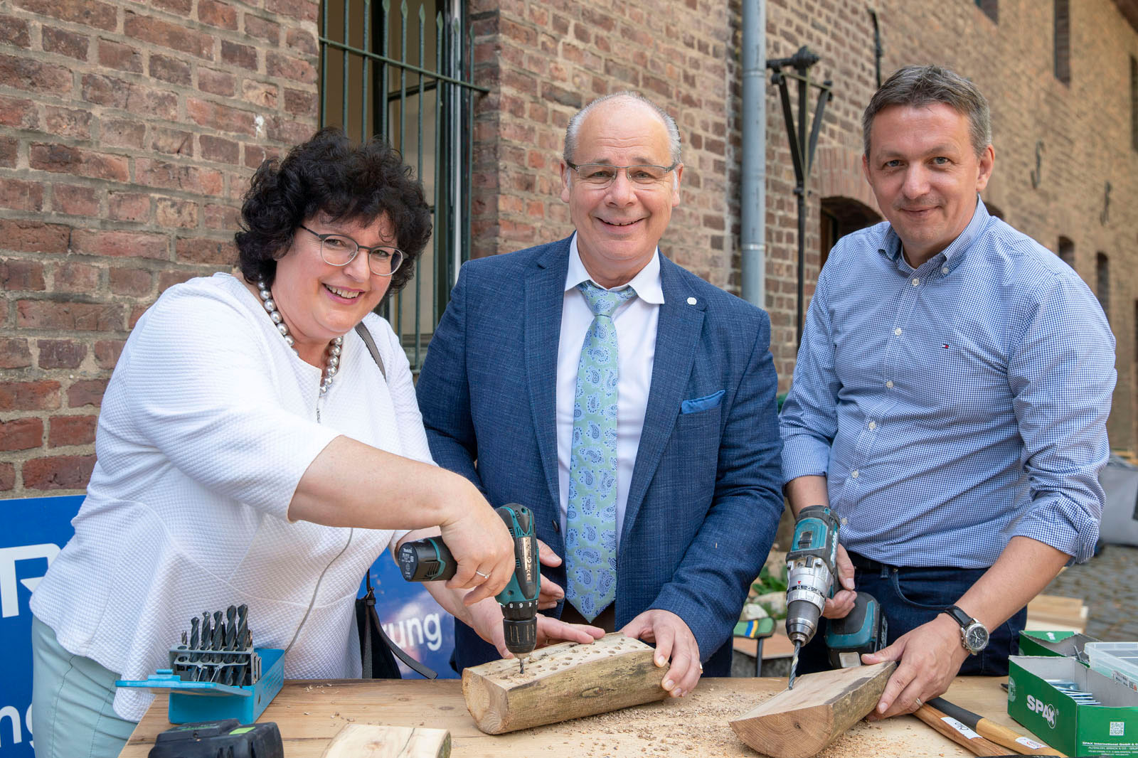 Drei Personen, die mit Holzarbeiten beschäftigt sind, verwenden Bohrmaschinen auf Holzplanken in der Nähe einer Backsteinmauer.