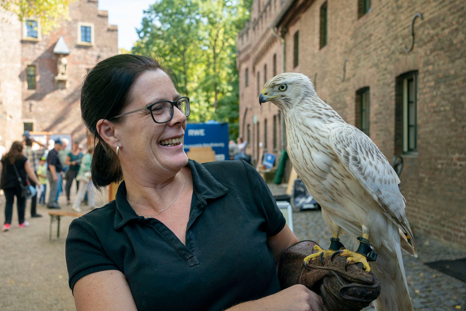 Eine Frau in einem schwarzen Shirt hält einen hellgefärbten Greifvogel auf ihrem Handschuh, umgeben von Menschen und Gebäuden.