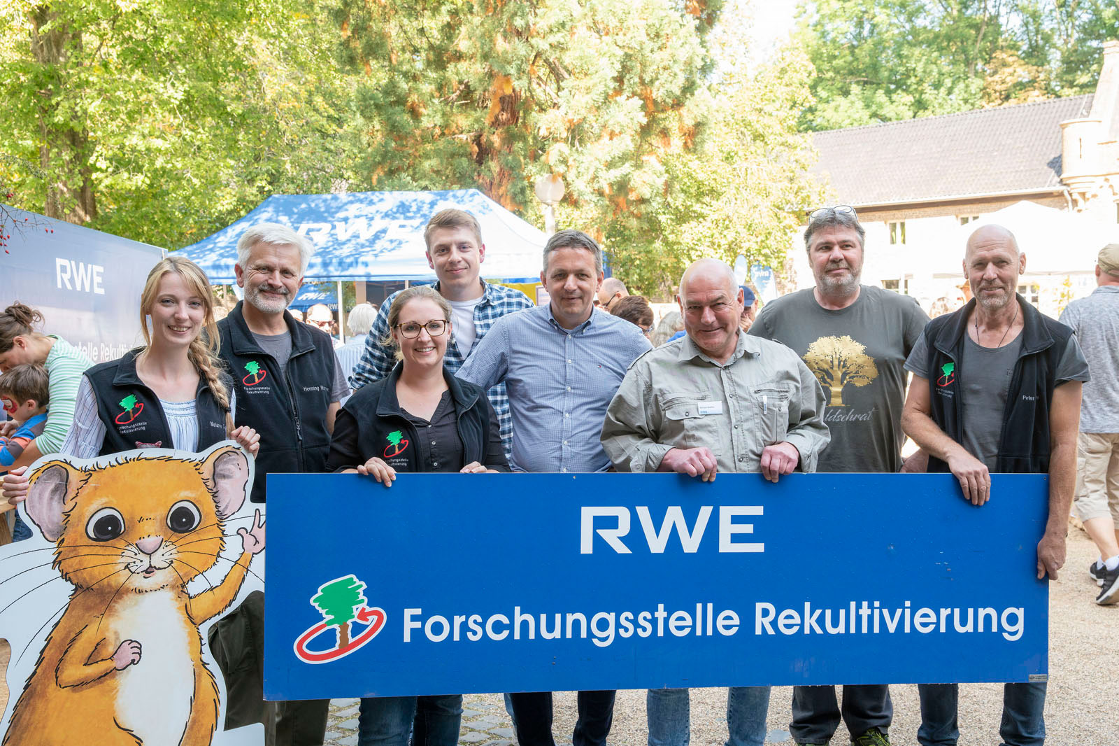 Eine Gruppe von Personen hält ein Schild für die Forschungsstelle Rekultivierung von RWE, mit einer Cartoon-Mausfigur links.