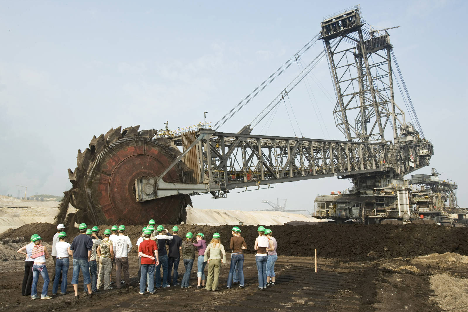 Ein großer Bagger arbeitet an einem Bergwerksstandort, während eine Gruppe von Personen mit grünen Helmen die Maschine beobachtet.