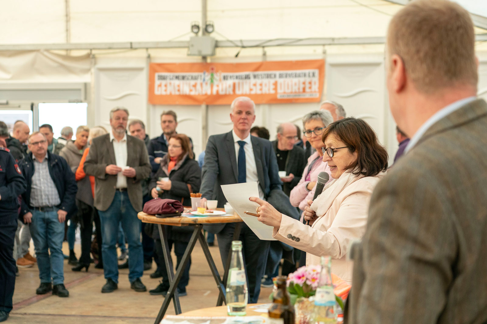Eine Menschenmenge versammelt sich drinnen bei einer Veranstaltung, mit einer Frau im Vordergrund, die spricht, und einem Banner im Hintergrund.