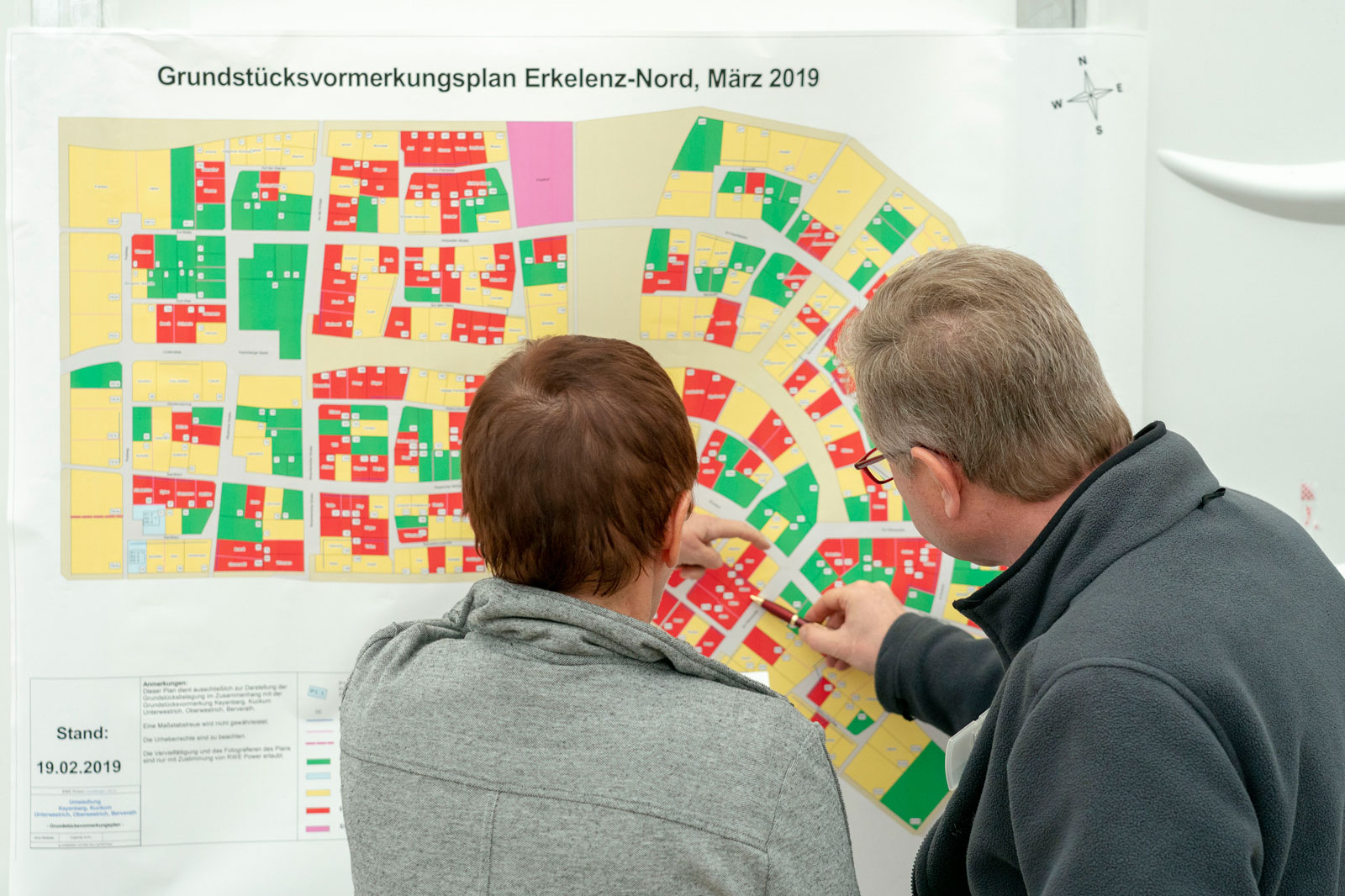 Zwei Personen betrachten einen Grundstücksplan mit farbigen Markierungen auf einer Wandtafel im Jahr 2019.