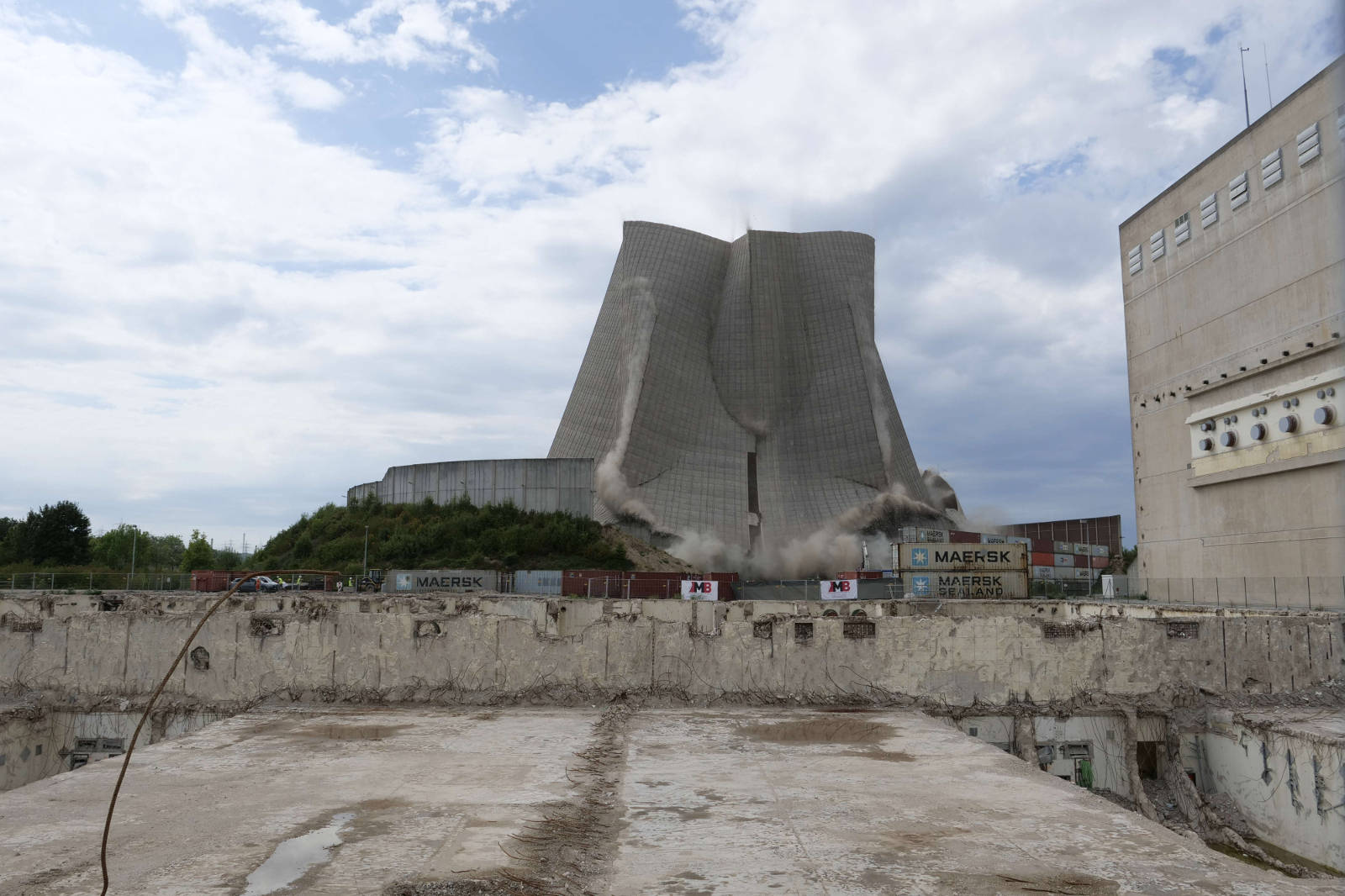 Ein Kühlturm eines Kernkraftwerks wird abgerissen, mit Trümmern und Versandcontainern im Vordergrund.