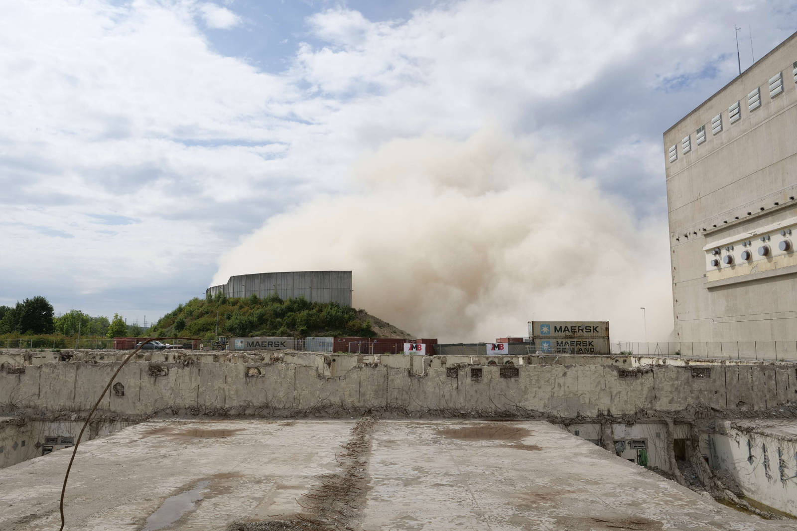 Eine Ansicht einer Abrissstelle mit Staubwolken von einer Explosion und Versandcontainern im Vordergrund.