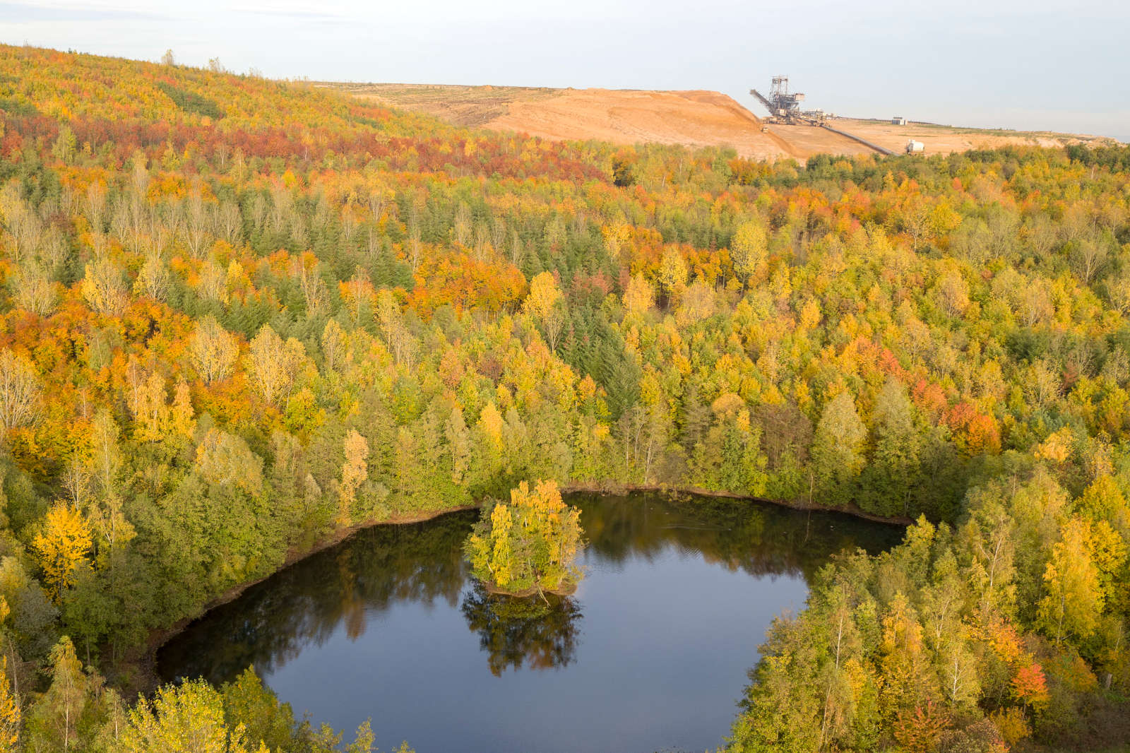 Eine lebhafte Herbstlandschaft mit einem kleinen See, umgeben von bunten Bäumen und einem Bergbaubetrieb im Hintergrund.