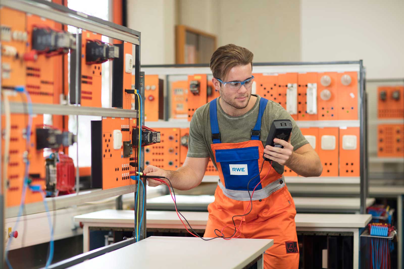 Ein Techniker in orangefarbenen Overalls misst elektrische Verbindungen mit einem Multimeter in einem Klassenraum mit orangen Schaltkreisen.