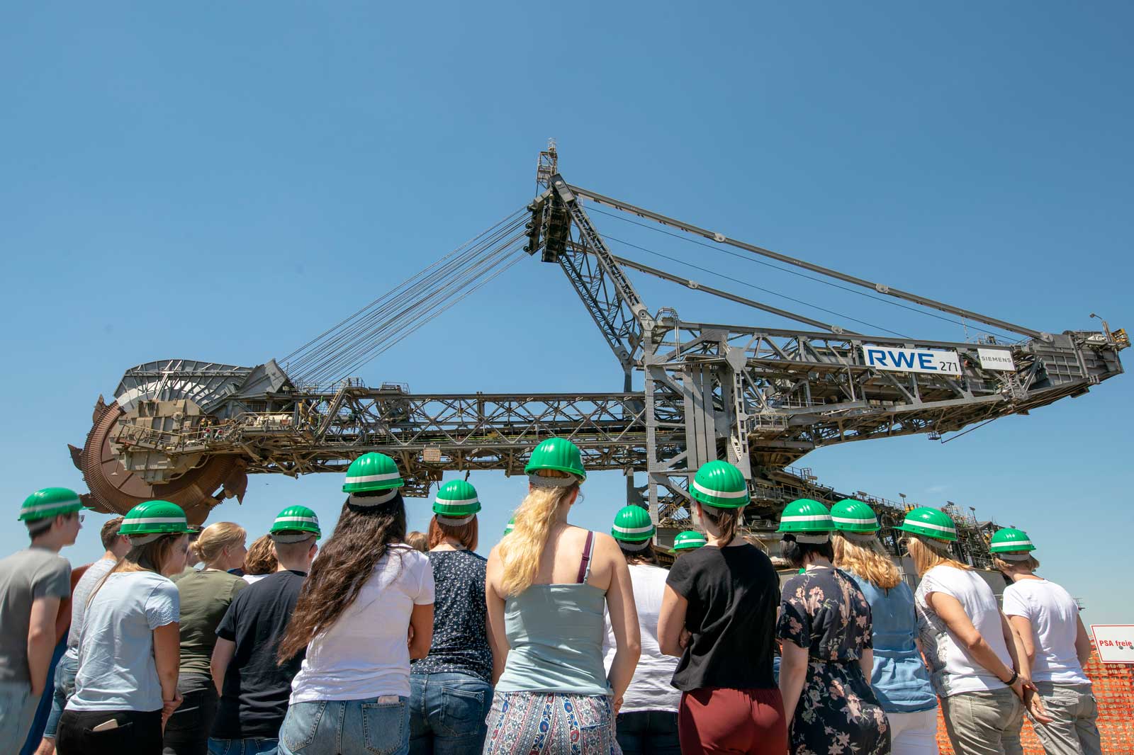 Eine Gruppe von Personen in grünen Helmen betrachtet einen großen industriellen Bagger vor einem klaren blauen Himmel.
