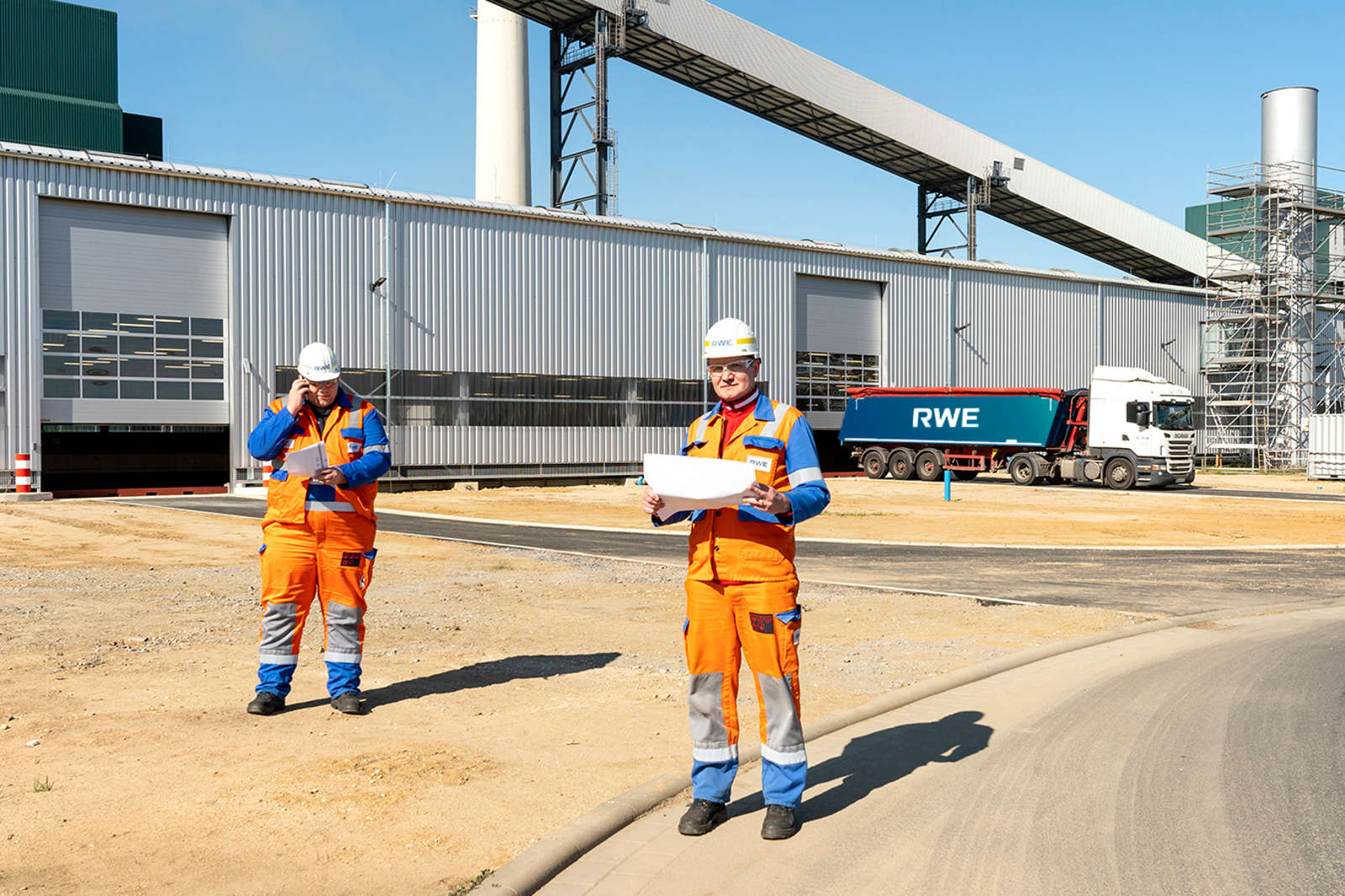 Zwei Bauarbeiter in orange und blauer Sicherheitskleidung besprechen Pläne vor einem großen Lagerhaus.