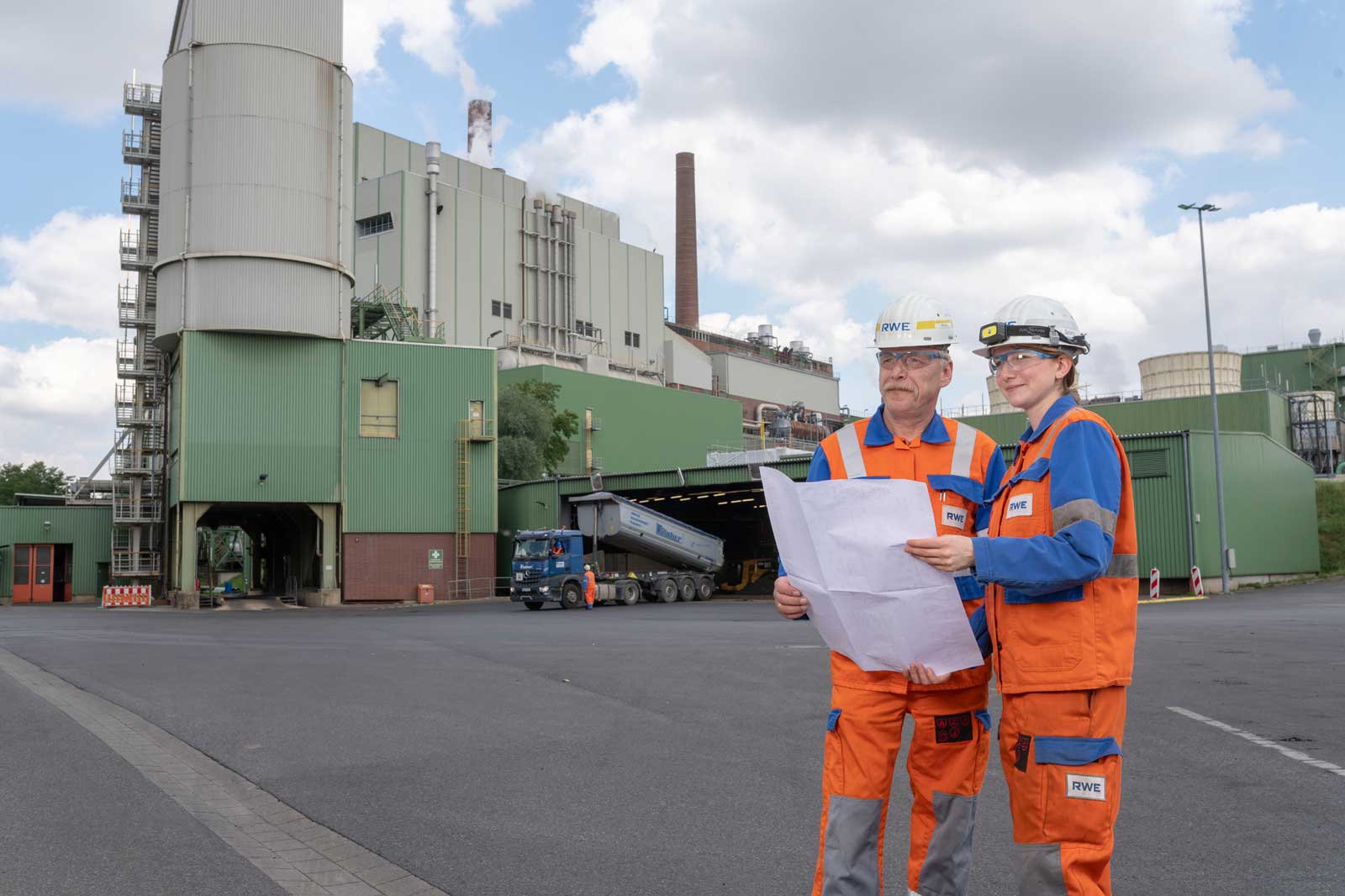 Zwei Arbeiter in orange-blauen Uniformen prüfen Pläne vor einem großen Industriegebäude unter einem bewölkten Himmel.