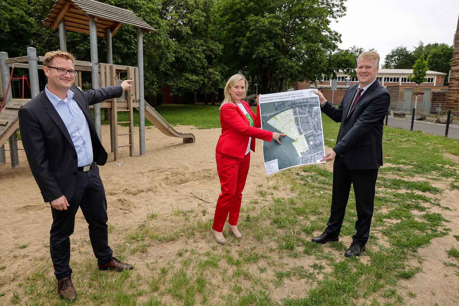 Drei Personen präsentieren eine Karte auf einem Spielplatz, umgeben von einem Holzspielgerät und grüner Landschaft.