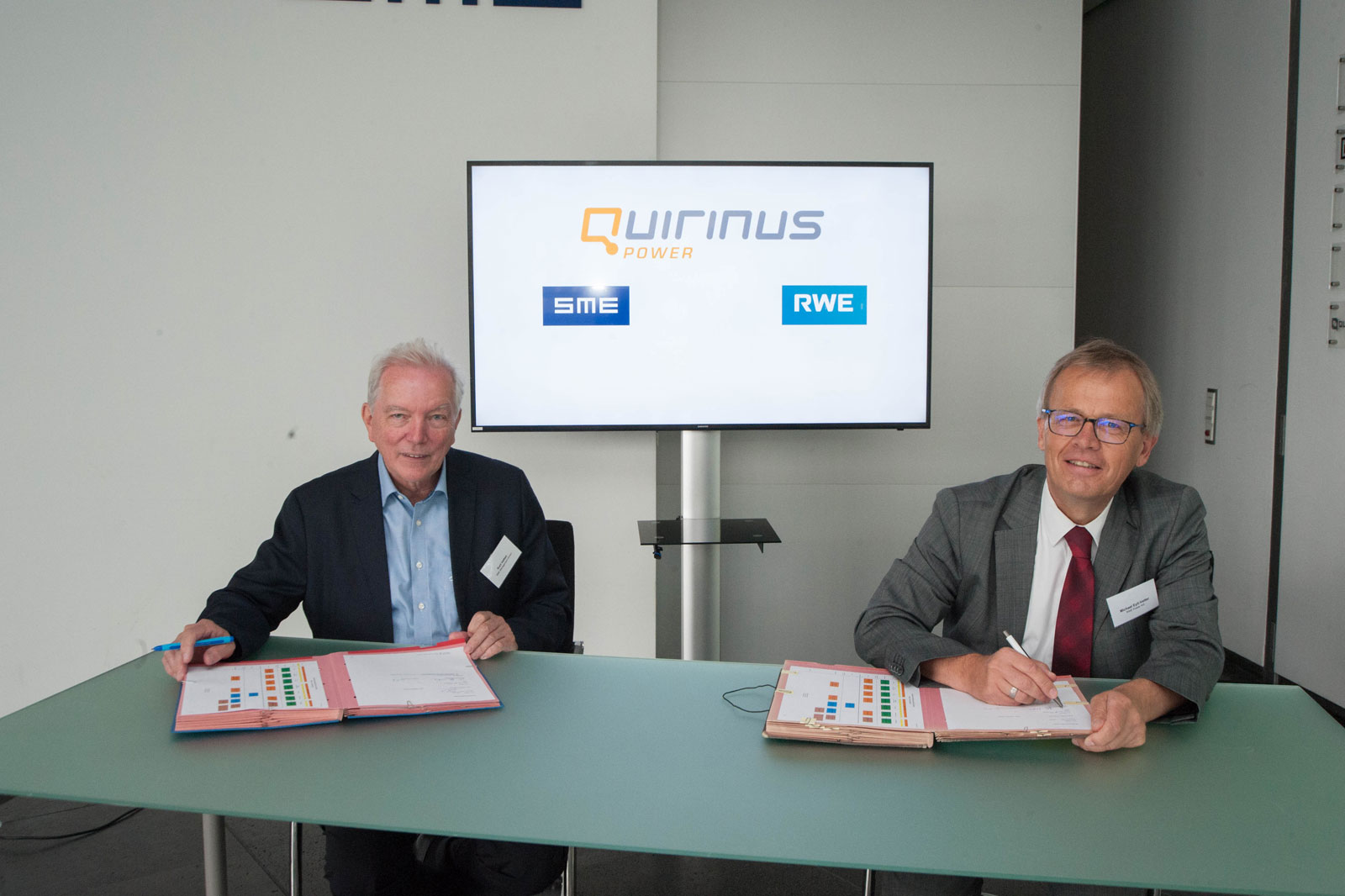 Zwei Männer sitzen an einem Tisch und unterzeichnen Dokumente, während ein Bildschirm Quirinus Power, SME und RWE im Hintergrund zeigt.