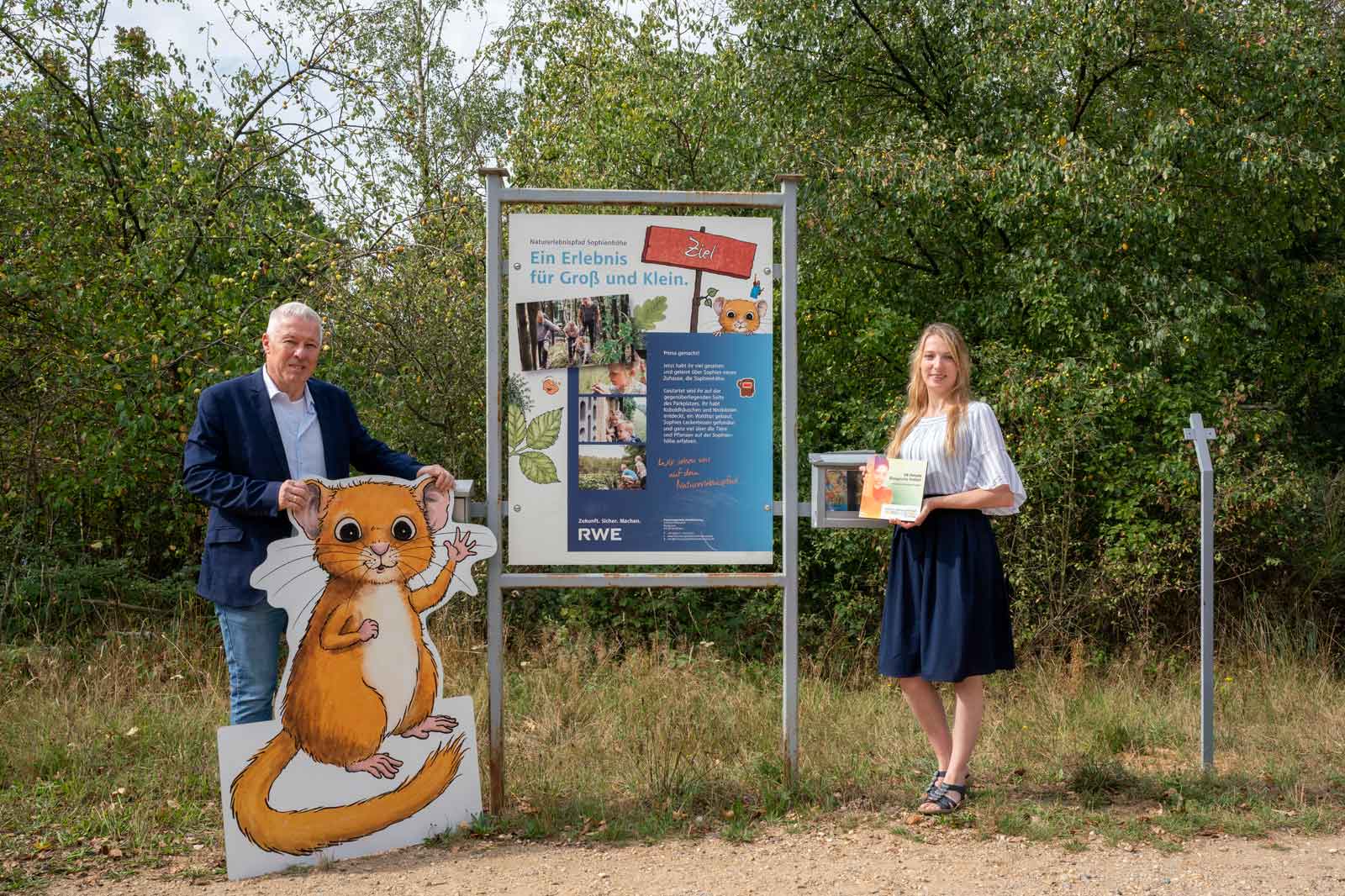 Zwei Personen stehen neben einem Schild, das eine Outdoor-Erfahrung bewirbt, mit einem Cartoon-Tier und informativem Text.