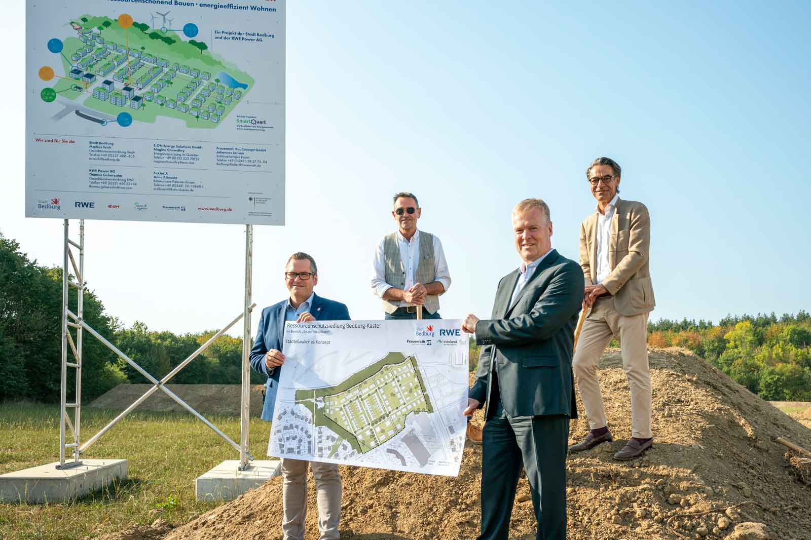 Vier Personen stehen auf einem Hügel und präsentieren ein Bauplan-Poster, während im Hintergrund ein Projektschild sichtbar ist.