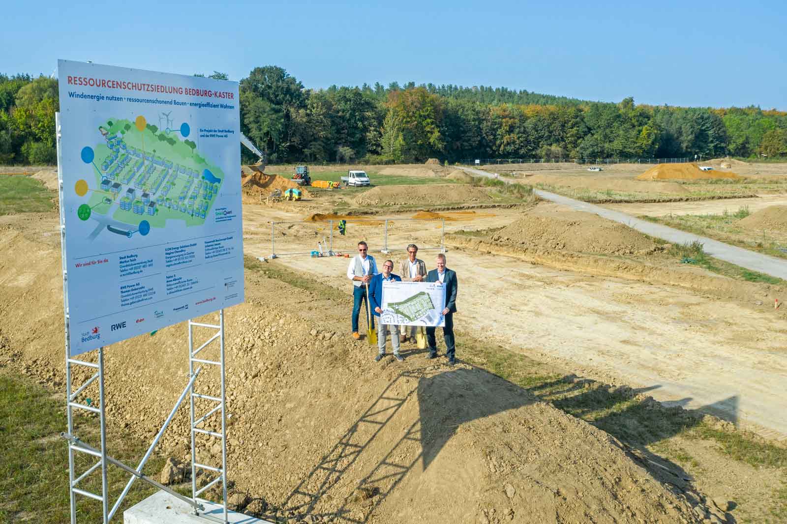 Bauplatz mit einem Projektplan, auf dem Personen eine Karte halten, umgeben von Erdarbeiten und Grünflächen.