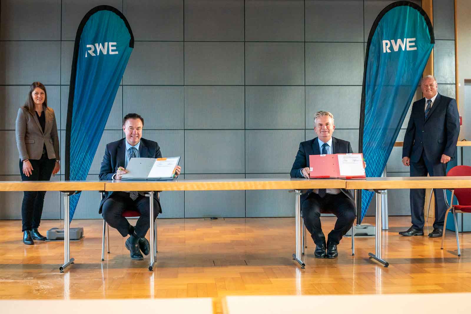 Zwei Männer sitzen an einem Holztisch mit Ordnern, RWE-Banner im Hintergrund, eine Frau steht in der Nähe.