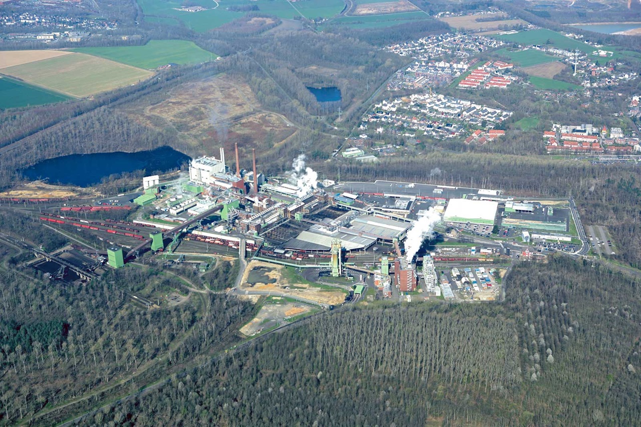 Luftaufnahme eines Industriekomplexes mit Rauchschächten, umgeben von Feldern, einem Wald und einem Wohngebiet.
