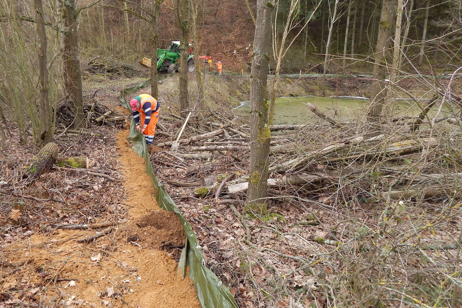 Ein Arbeiter in einer orangefarbenen Uniform verlegt eine grüne Plane in einem Waldgebiet, während im Hintergrund Maschinen und Kollegen sichtbar sind.