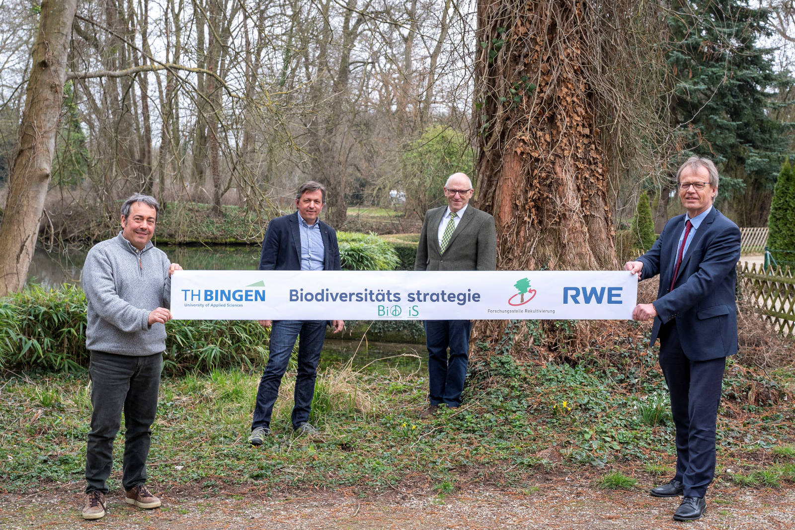 Vier Personen halten ein Banner zur Förderung der Biodiversitätsstrategie in einem bewaldeten Gebiet mit einem Teich im Hintergrund.