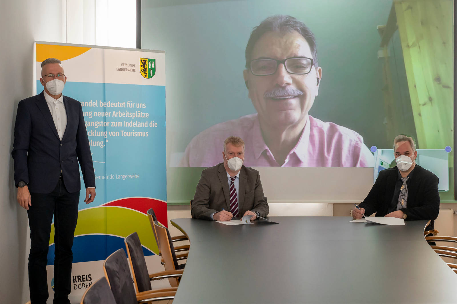 Eine Gruppenbesprechung mit drei Teilnehmern, zwei persönlich und einer auf dem Bildschirm, neben einem Werbebanner.