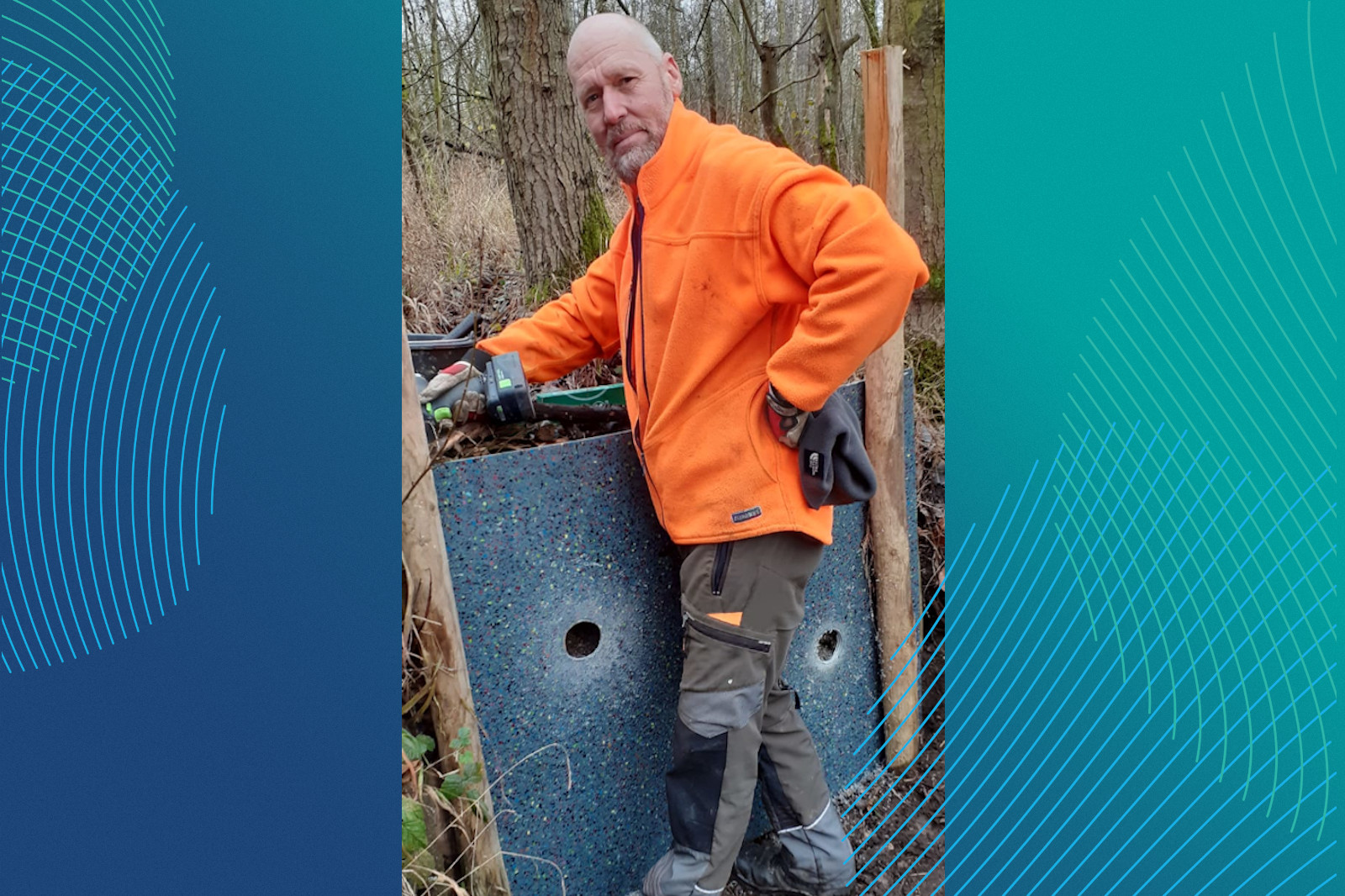 Eine Person in einer orangefarbenen Jacke steht neben einem bunten Panel in einem Waldgebiet und hält ein Werkzeug in der Hand.