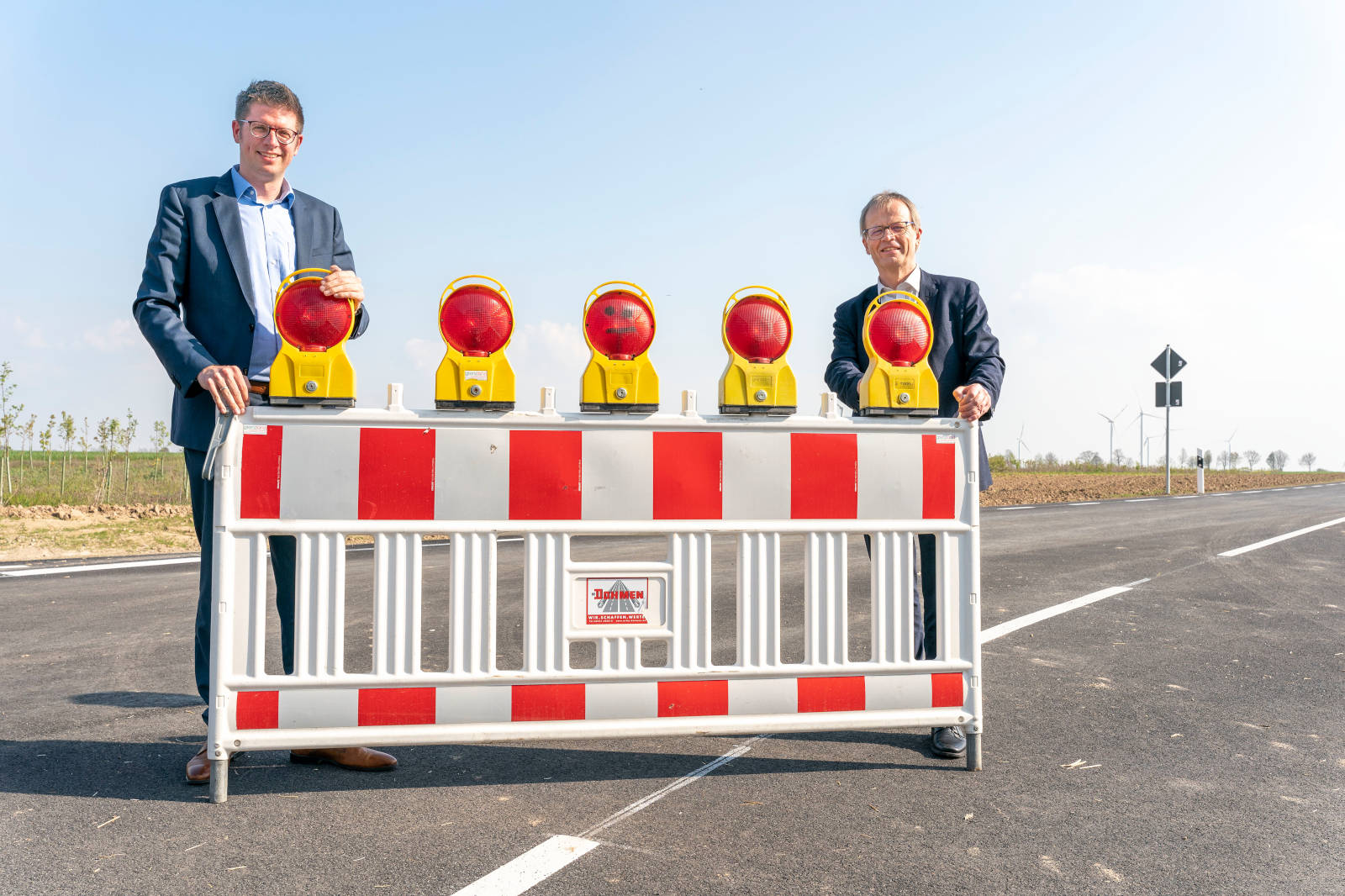 Zwei Männer in Anzügen stehen hinter einer Verkehrssperre mit gelben und roten Warnlichtern vor einem klaren Himmel.