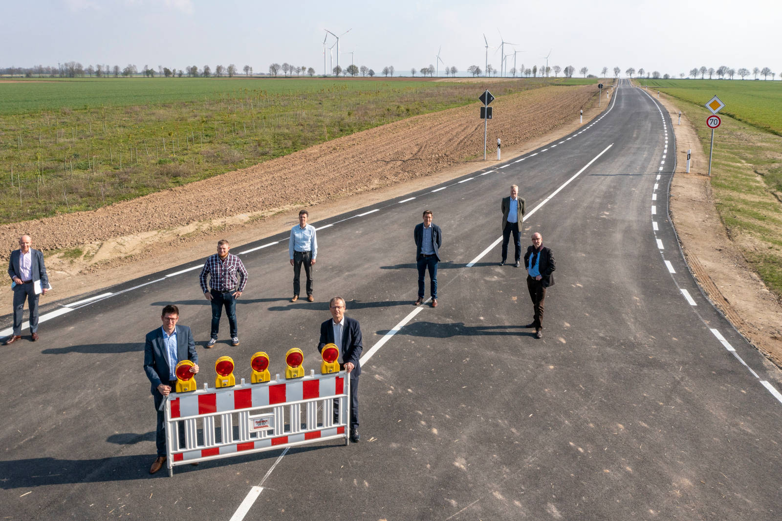 Eine Gruppe von Menschen steht auf einer neuen Straße mit einer Barriere und Verkehrslichtern, umgeben von Feldern und Windkraftanlagen.