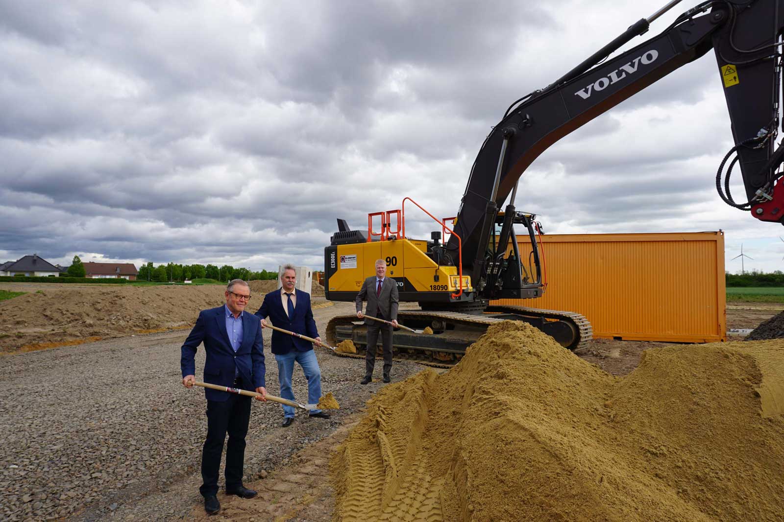 Drei Männer in formeller Kleidung führen eine Grundsteinlegung mit einem gelben Bagger und einem Sandhaufen durch.