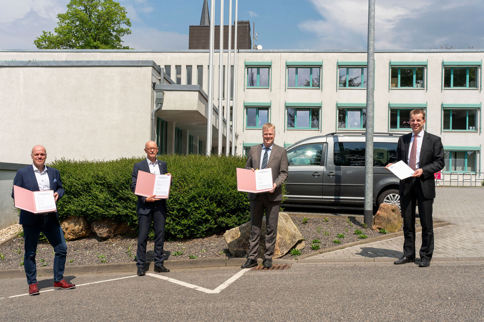 Vier Personen stehen vor einem modernen Gebäude und halten pinkfarbene Urkunden unter einem sonnigen Himmel.