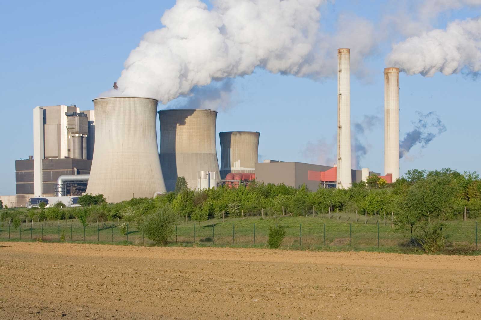 Ein Kraftwerk mit Kühltürmen und Schornsteinen, die Dampf und Rauch ausstoßen, umgeben von Grünflächen und Ackerland.