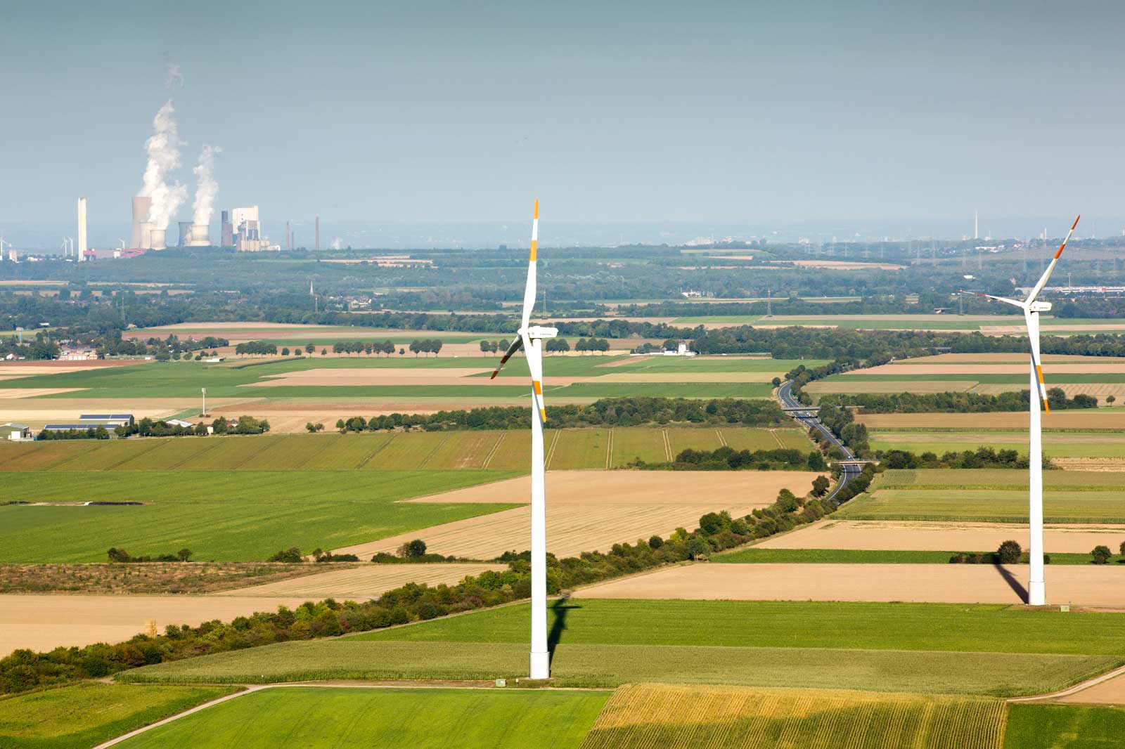 Eine Sicht auf Windkraftanlagen in einer grünen Landschaft mit Feldern und Fabriken, die Rauch im Hintergrund ausstoßen.