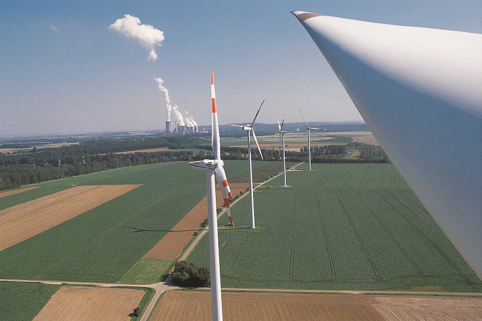 Luftaufnahme von Windkraftanlagen in einem Feld mit einem rauchenden Kraftwerk im Hintergrund.