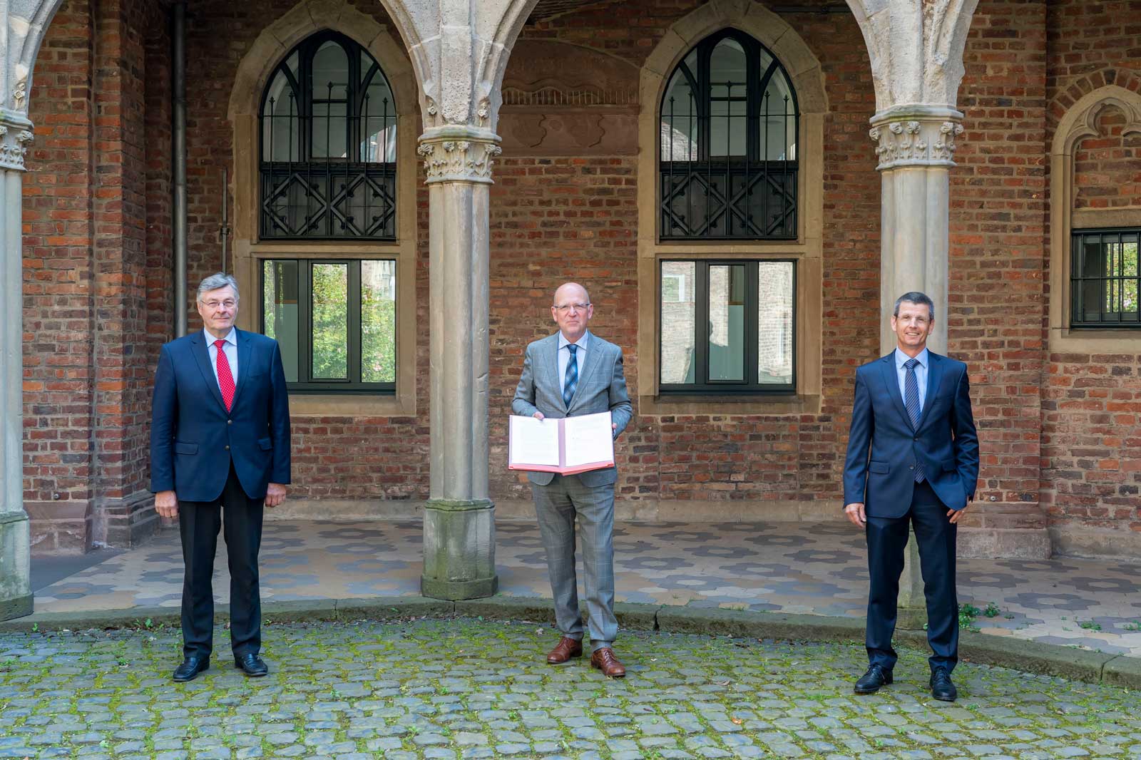 Drei Männer stehen in einem Innenhof mit Backsteinwänden, einer hält ein Dokument. Sie sind formell gekleidet.