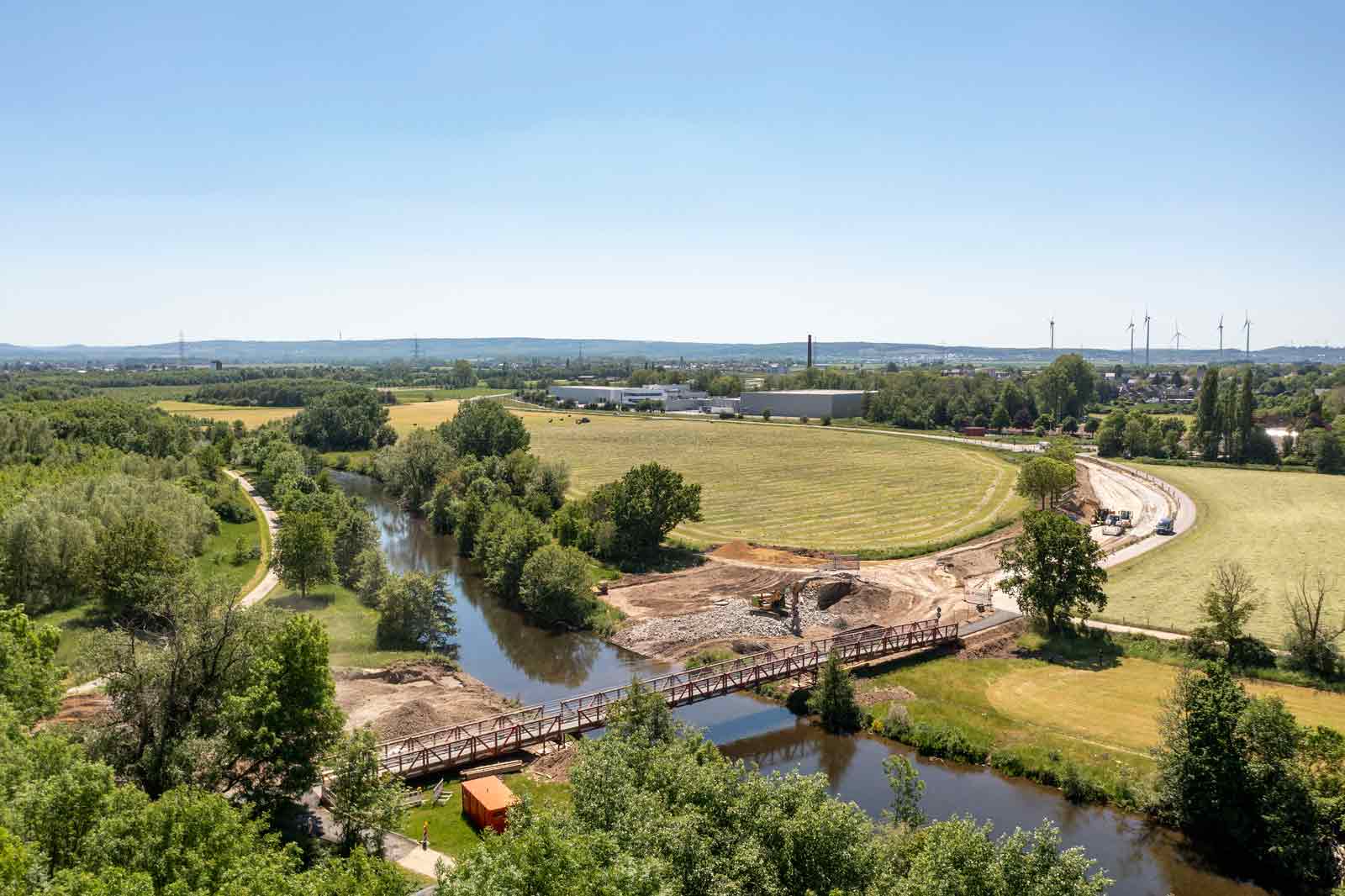 Eine malerische Aussicht auf ein Flussufer mit einer Brücke, umgeben von üppigem Grün und Feldern unter einem klaren blauen Himmel.