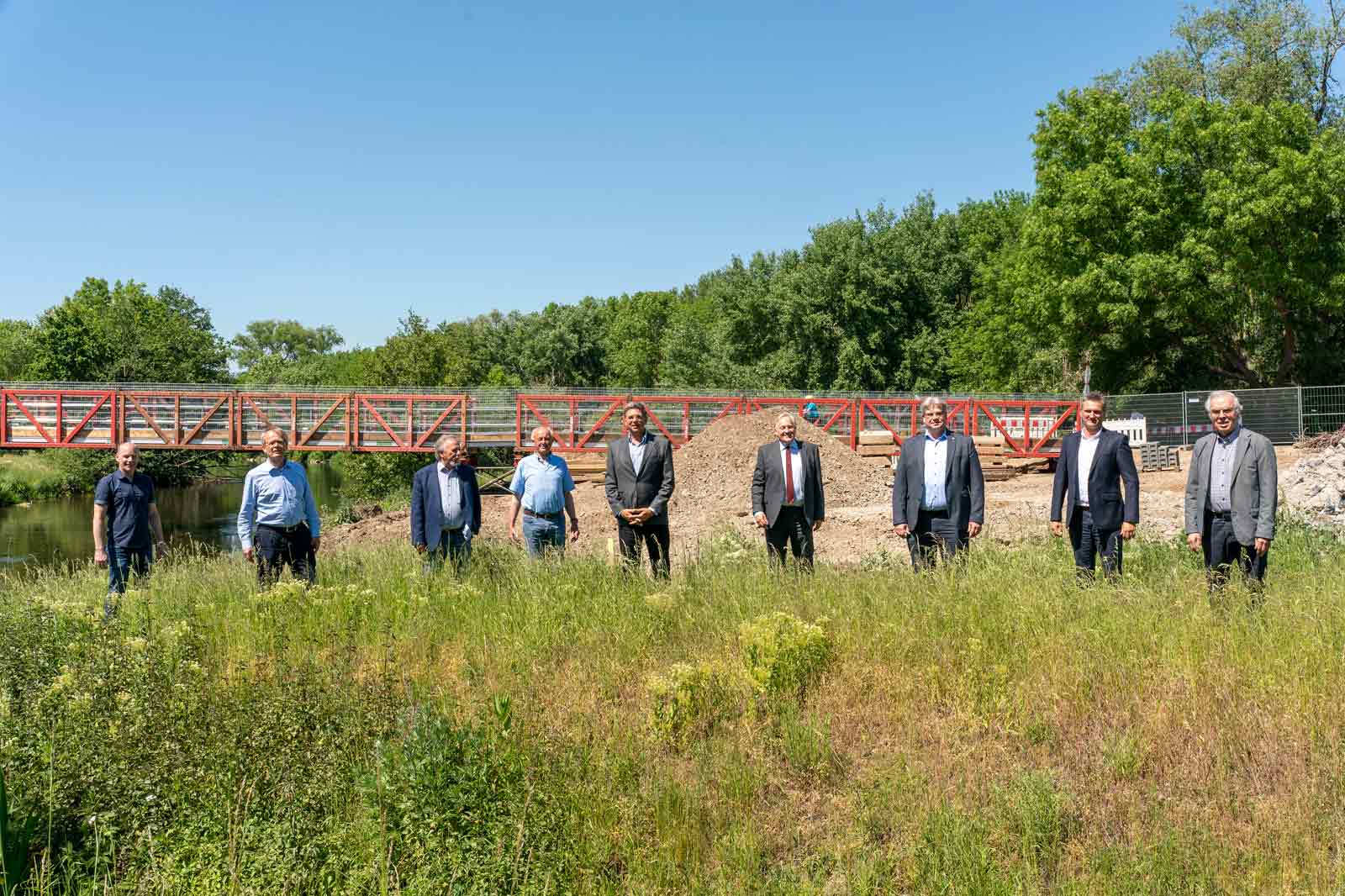 Eine Gruppe von Menschen in formeller Kleidung steht auf einem grasbewachsenen Gelände in der Nähe einer roten Fußgängerbrücke, umgeben von Bäumen.