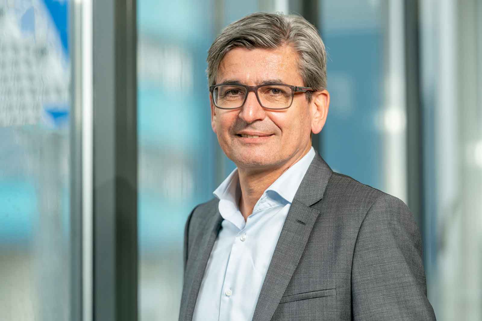 Ein Mann in einem grauen Anzug und einem weißen Hemd steht neben großen Fensterfronten in einer modernen Büroumgebung.