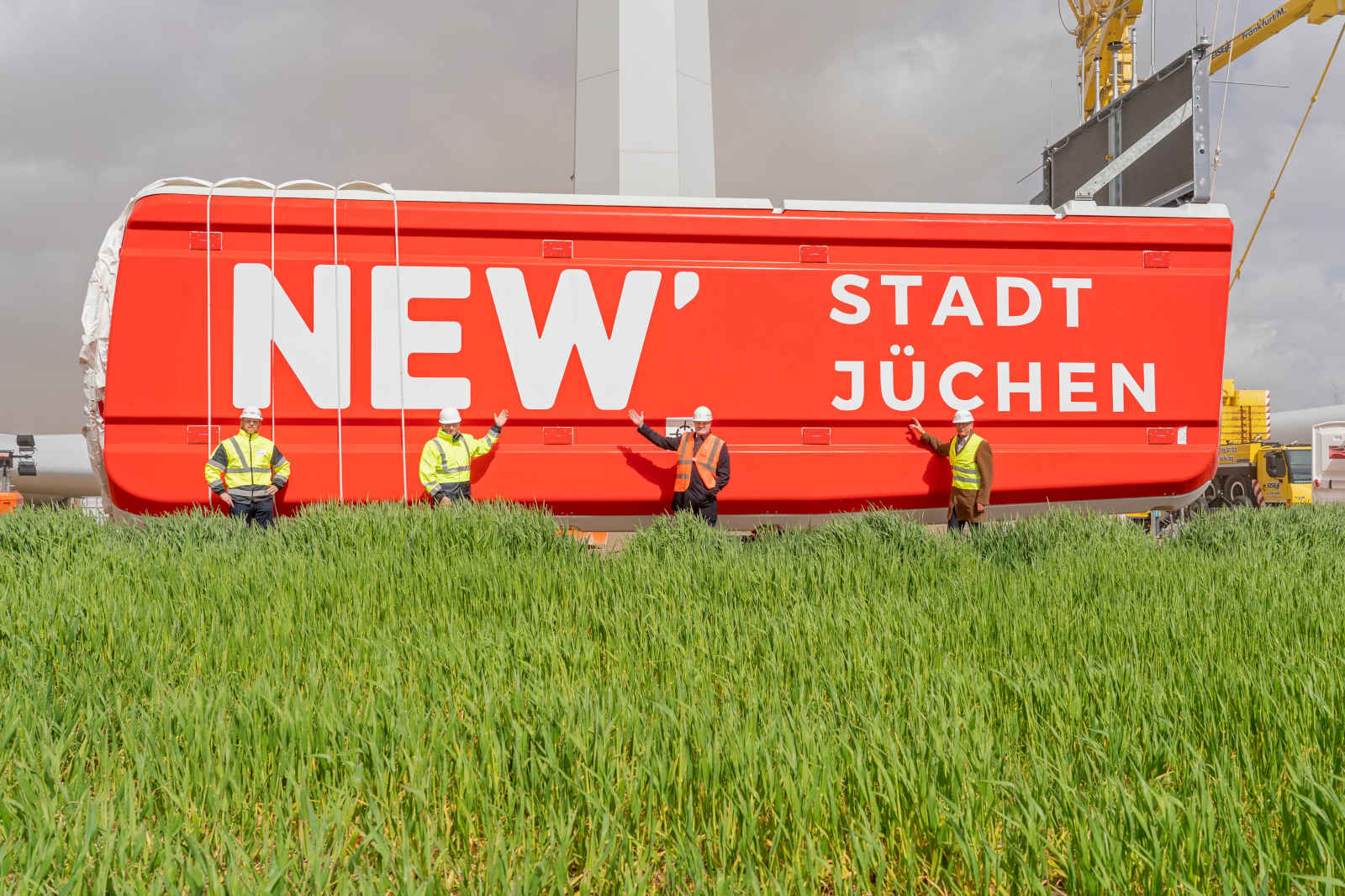 Ein rotes Schild mit der Aufschrift 'NEW STADT JÜCHEN' und mehreren Sicherheitsmitarbeitern vor hohem, grünem Gras.