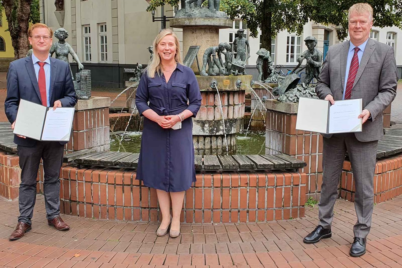 Drei Personen stehen neben einem Brunnen. Zwei halten Dokumente, während eine in einem blauen Kleid steht. Skulpturen sind im Hintergrund.