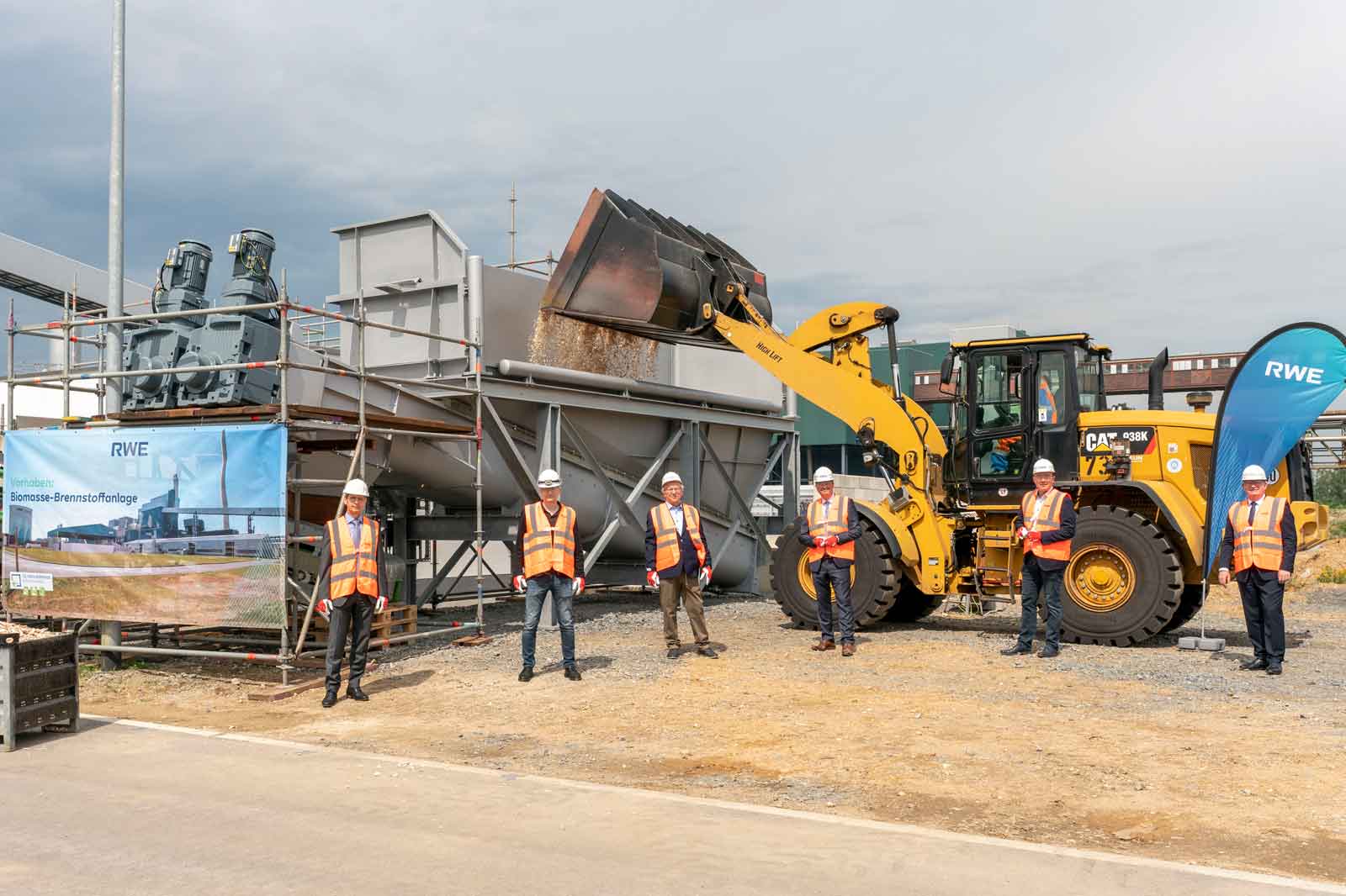 Ein gelber Lader entleert Material in eine graue Biomasseanlage, mit Arbeitern in Sicherheitskleidung und einem RWE-Banner im Hintergrund.
