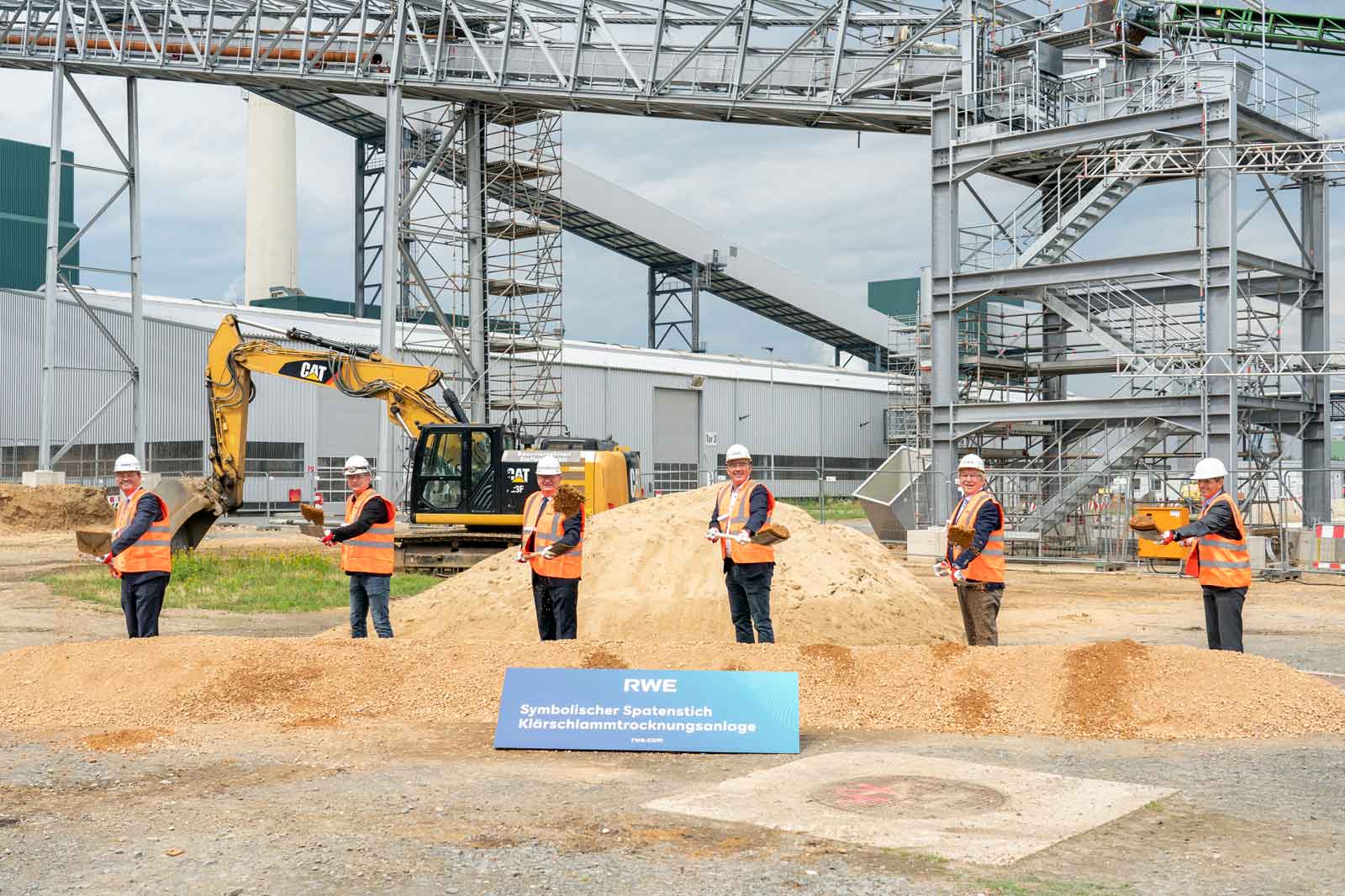 Fünf Männer in Sicherheitswesten stehen um einen Sandhaufen. Im Hintergrund ist ein Bagger und eine industrielle Anlage zu sehen.