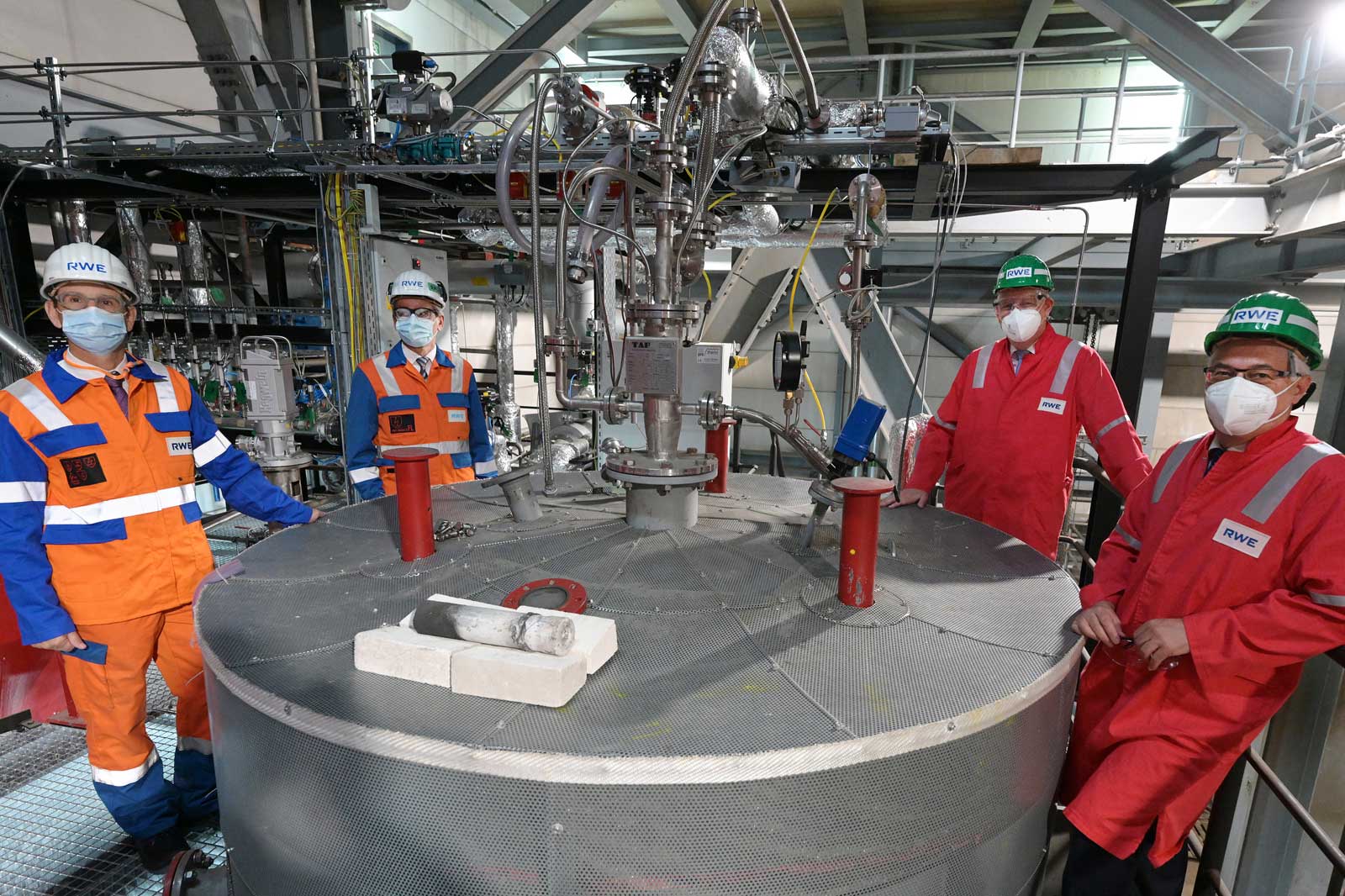Vier Arbeiter in Schutzausrüstung stehen in einer Industrieumgebung neben einer großen kreisförmigen Maschinenplattform.