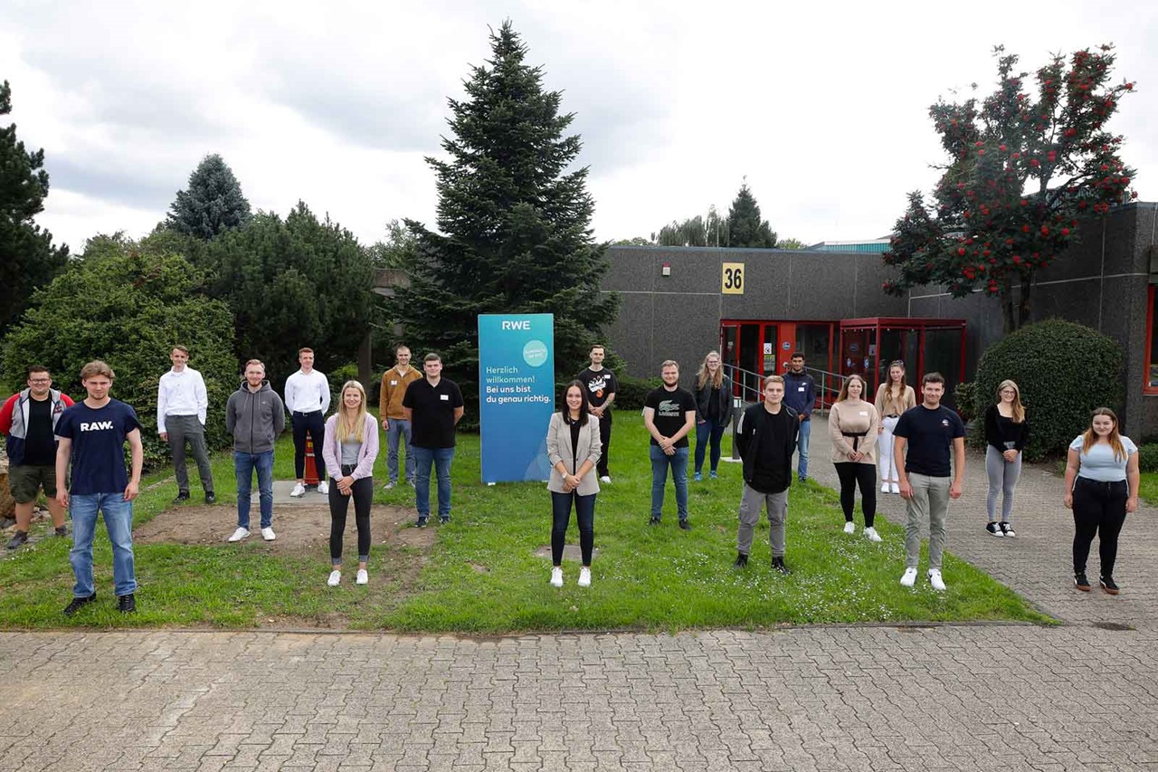 Eine Gruppe von Menschen steht im Freien vor einem Gebäude, umgeben von Grünflächen und einem Willkommensschild.